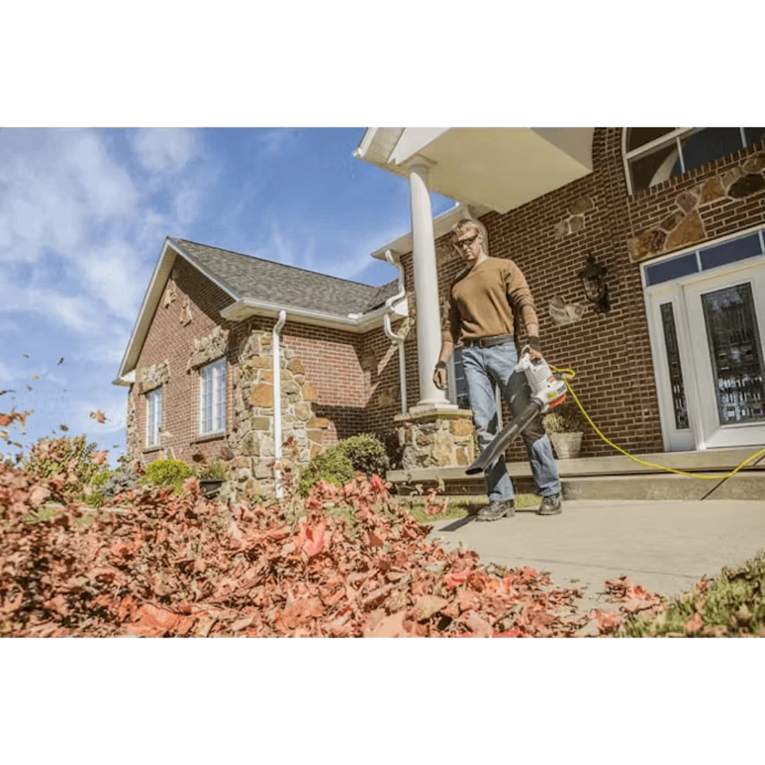 Man using an electric handheld blower on leaves outside a brick and stone house in a home improvement setting