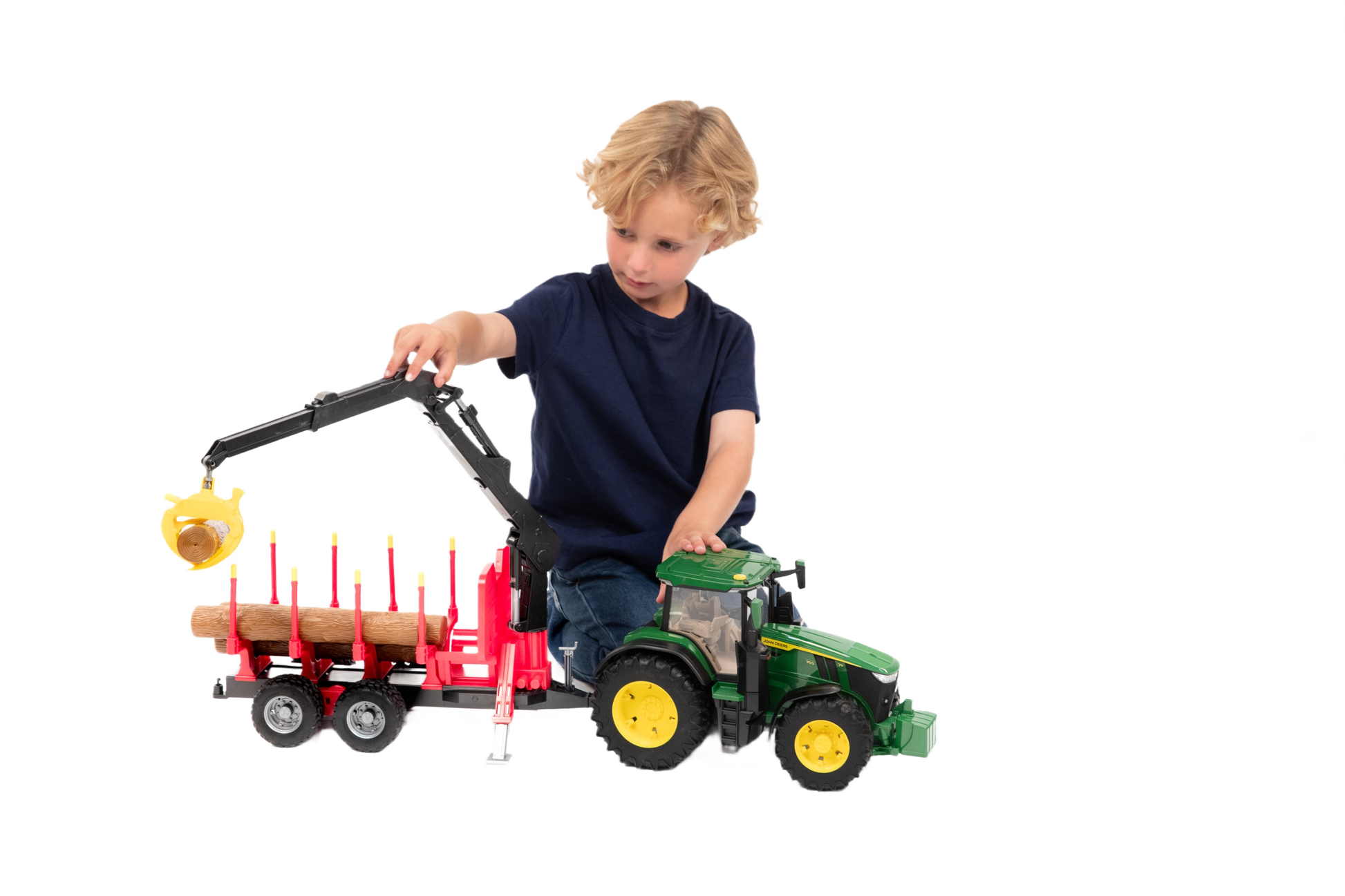 Young child playing with green and yellow Bruder tractor connected to red forestry trailer carrying wooden logs