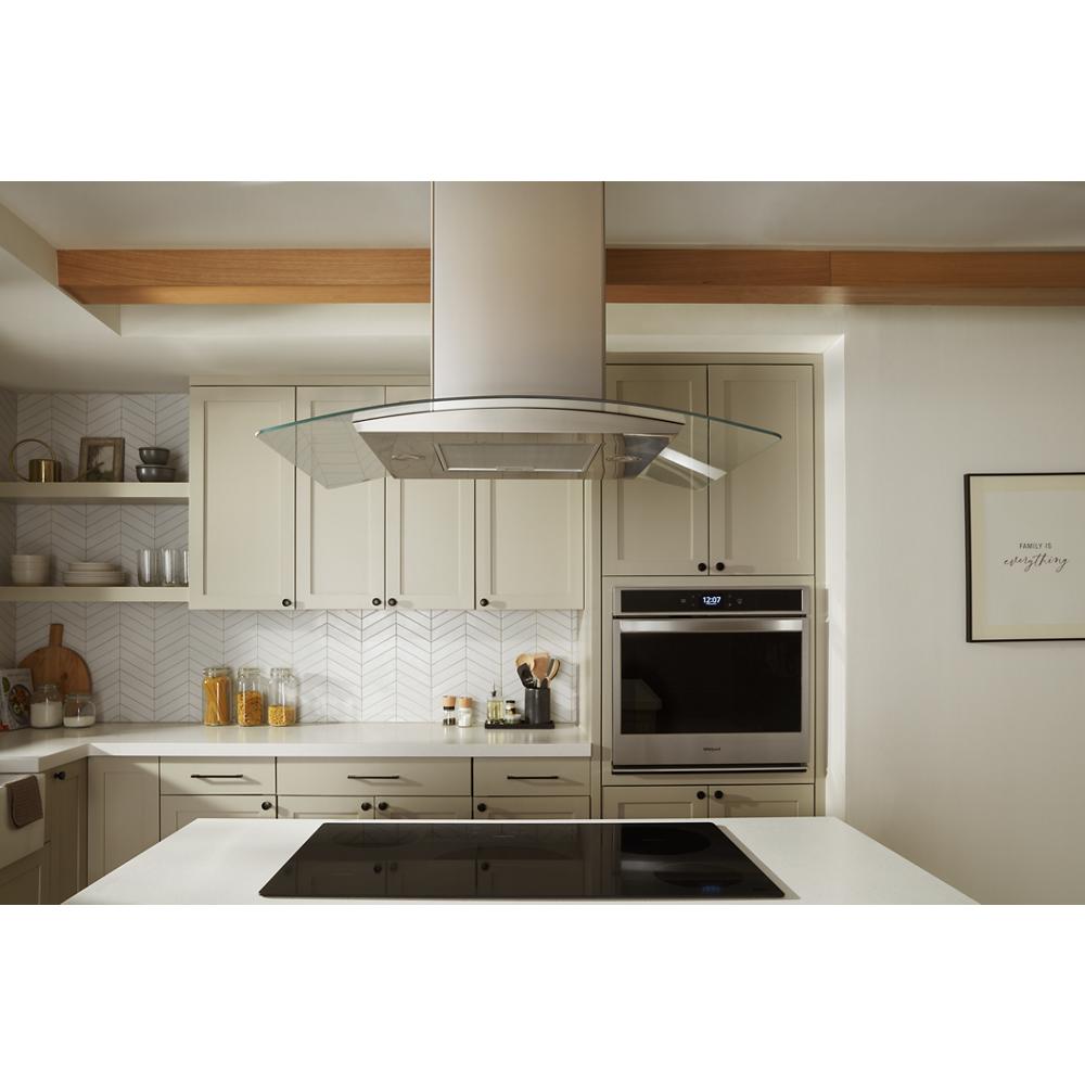 Modern kitchen with curved glass wall mount range hood above black cooktop and beige cabinetry in appliances collection