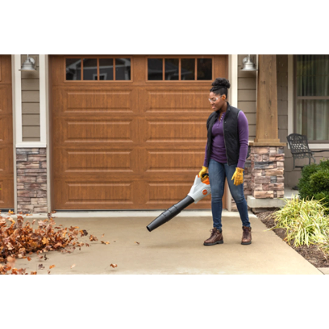 Person using cordless handheld leaf blower on driveway in front of residential garage door for home cleanup