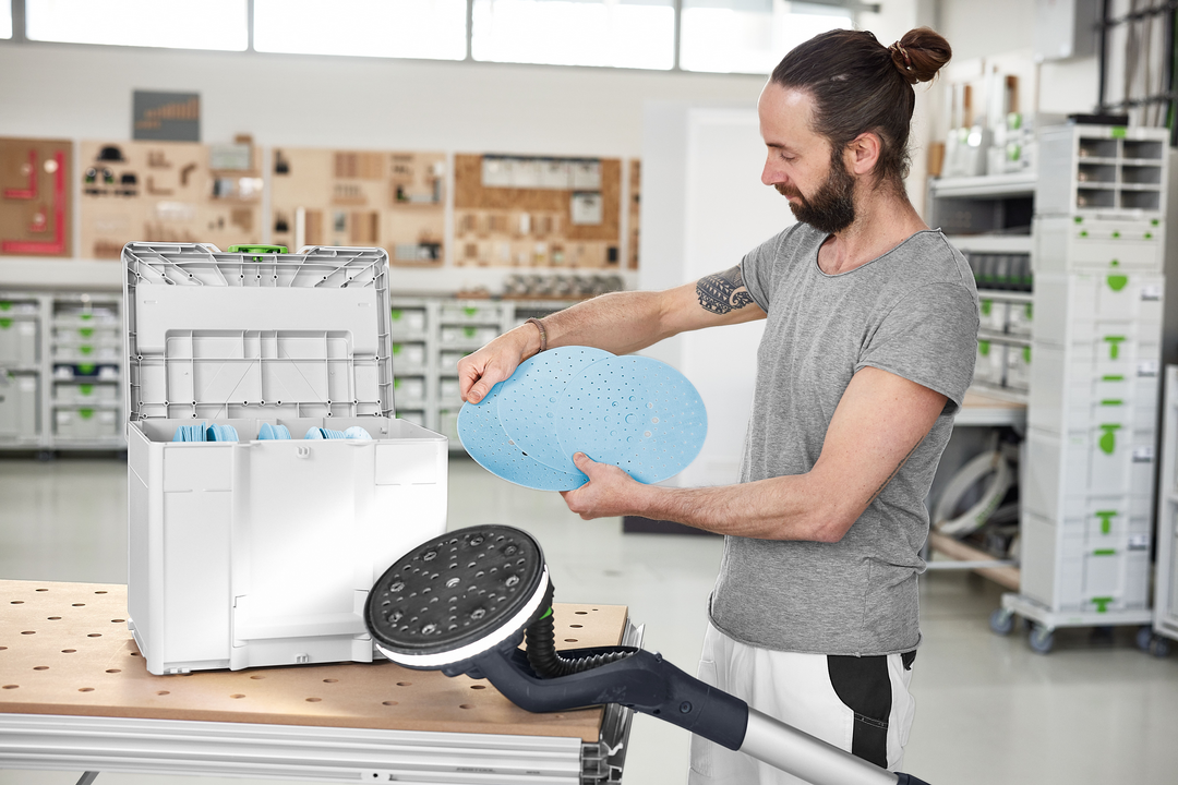 Person holding blue abrasive discs next to a white Festool Granat abrasives Systainer in workshop setting