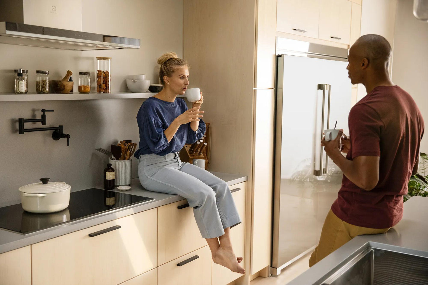Modern kitchen scene with a person sitting on light cabinetry near a sleek induction cooktop and another person standing by a refrigerator