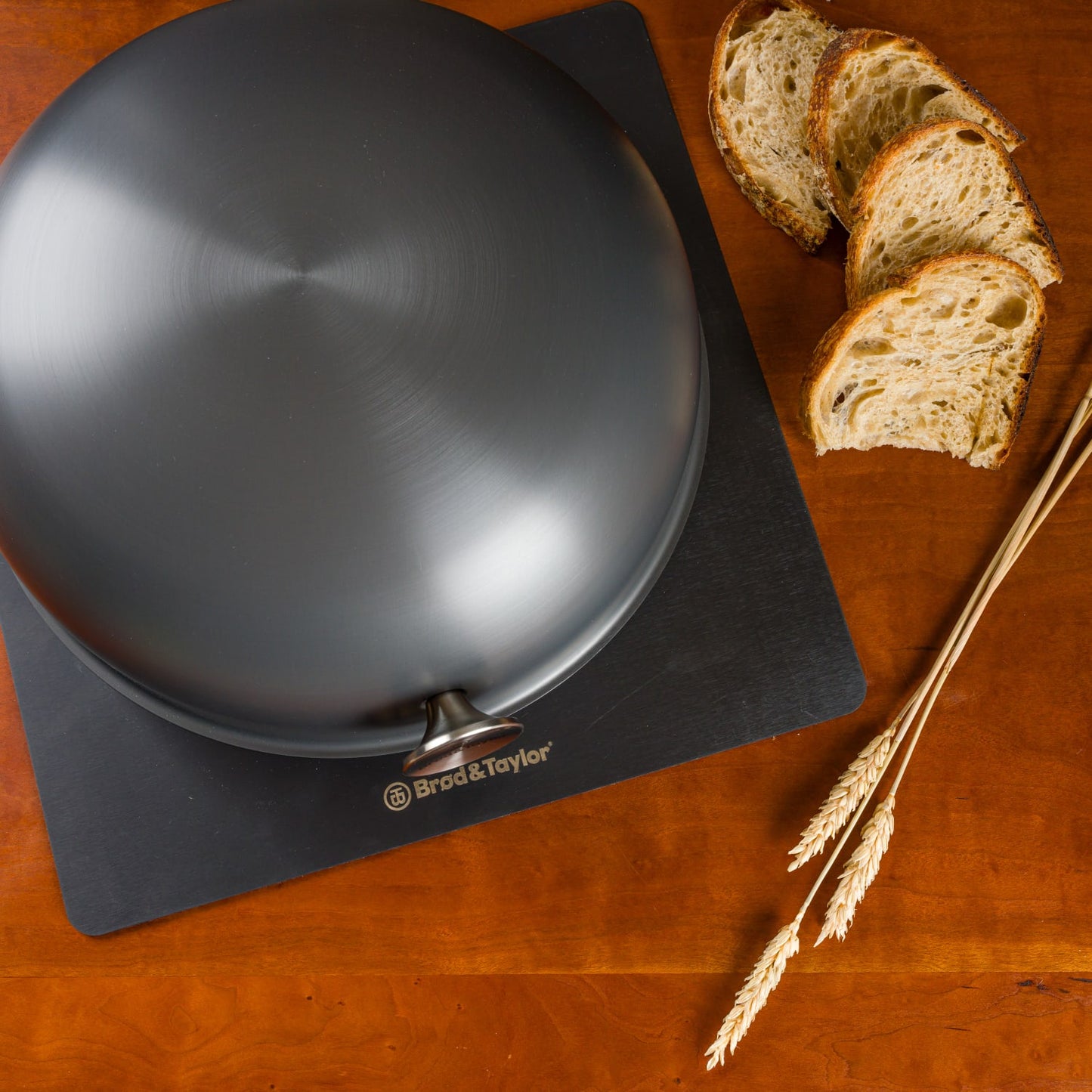 Matte black domed baking shell resting on branded board beside sliced artisan bread and wheat stalks in kitchenware collection