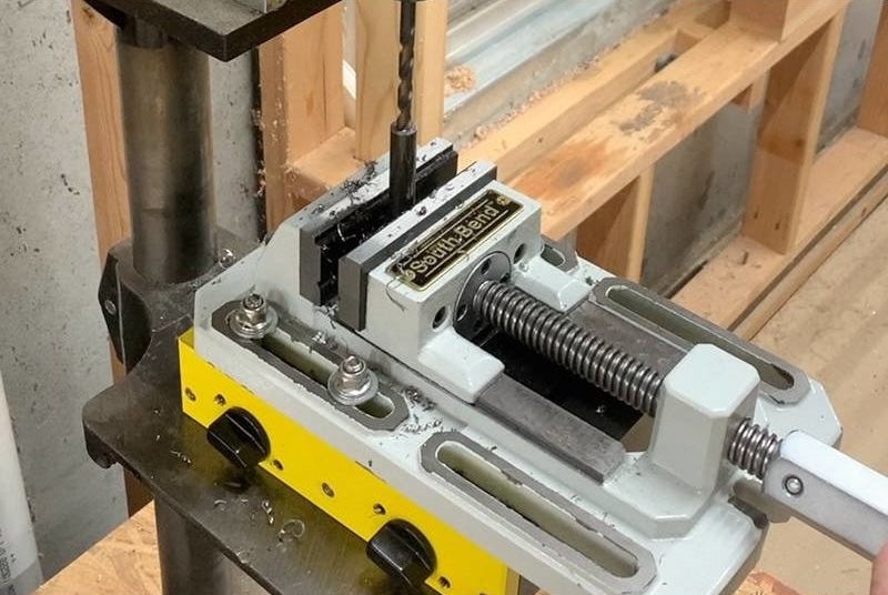 Magswitch drill press vise mount clamped on a metal press table with wood framing and drill bit in workshop setting