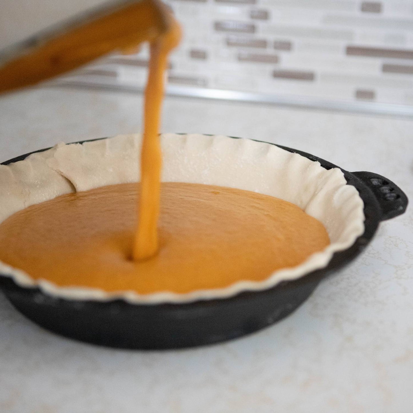 Pouring smooth pumpkin filling into seasoned cast iron pie pan with crimped edges on kitchen countertop