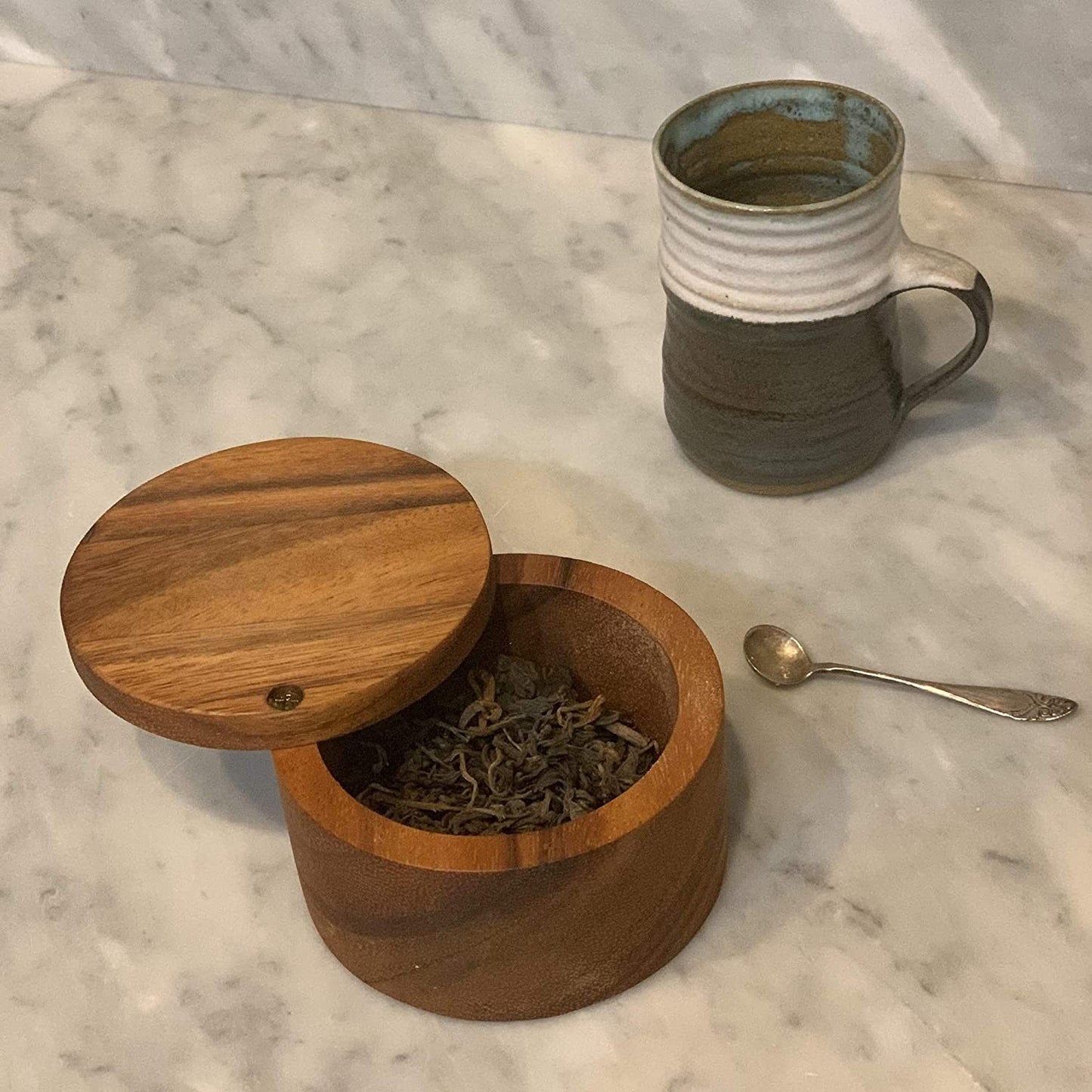 Acacia wood container with hinged lid storing dried herbs beside ceramic mug and small spoon in kitchenware setting