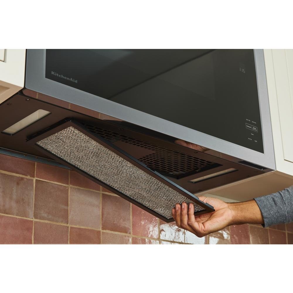Hand removing mesh filter from underside vent on stainless steel over-the-range microwave in kitchen setting