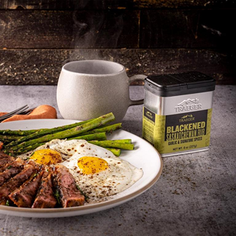 Tin of blackened Saskatchewan rub with cooked steak, asparagus, and fried eggs on a white plate in outdoor living setting