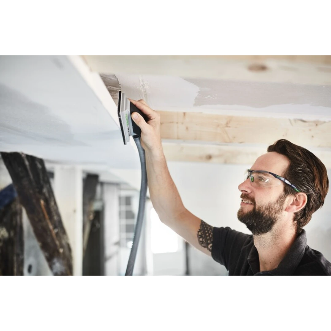 Man wearing safety glasses using hand sanding block with dust extraction for drywall finishing in home improvement project