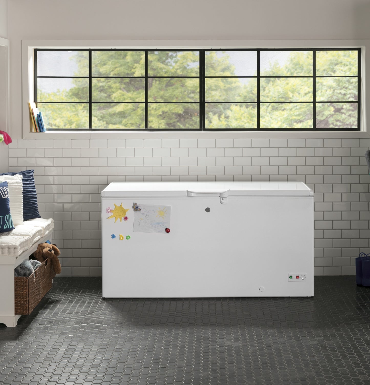 White manual defrost chest freezer with decorative magnets in a bright kitchen with subway tile backsplash and large window