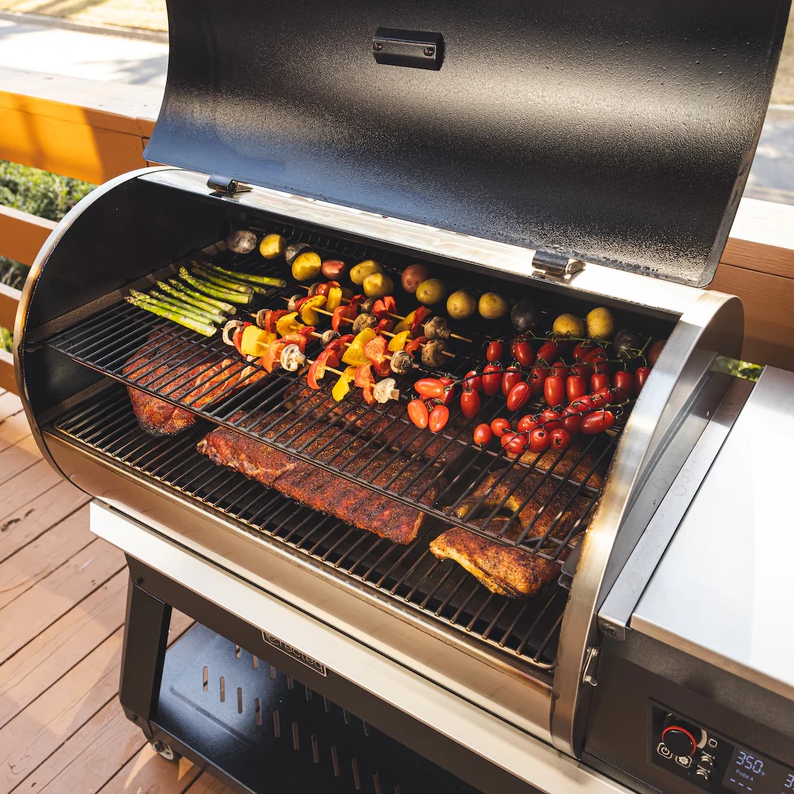 Stainless steel pellet grill open to reveal assorted grilled vegetables, skewers, ribs, and chicken on two cooking racks, ideal for outdoor cooking and grilling.