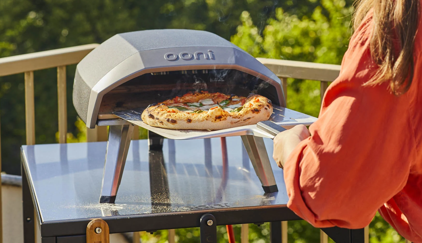Person using a black and silver ooni portable pizza oven on a metal cart in an outdoor living setting