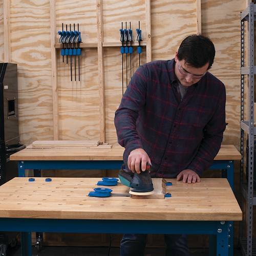 Man using power sander on hardwood bench top with dog holes in a woodworking shop for home improvement tools