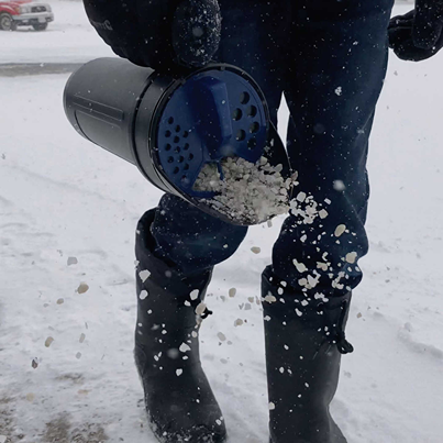 Black handheld spreader discharging salt onto snowy ground worn by person in black boots and winter clothing