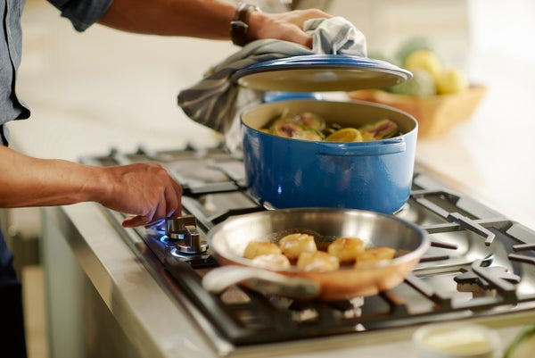 Person adjusting flame on stainless steel gas cooktop while cooking with blue pot and frying pan in kitchen setting