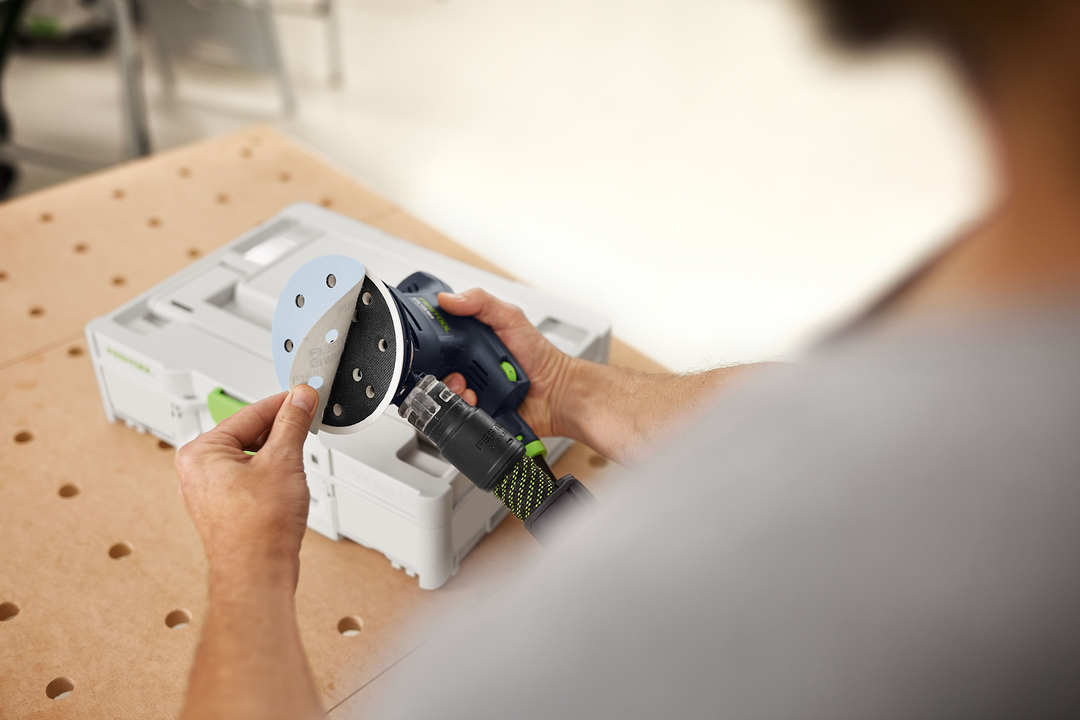 Person attaching gray sanding disc to Festool power sander with white storage case on workbench in workshop