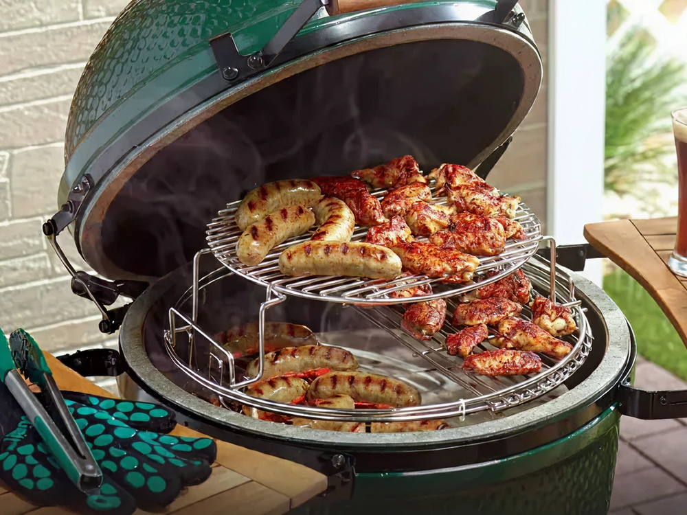 Green ceramic grill with two-tiered rack cooking grilled sausages and chicken in an outdoor patio area with barbecue tools nearby.