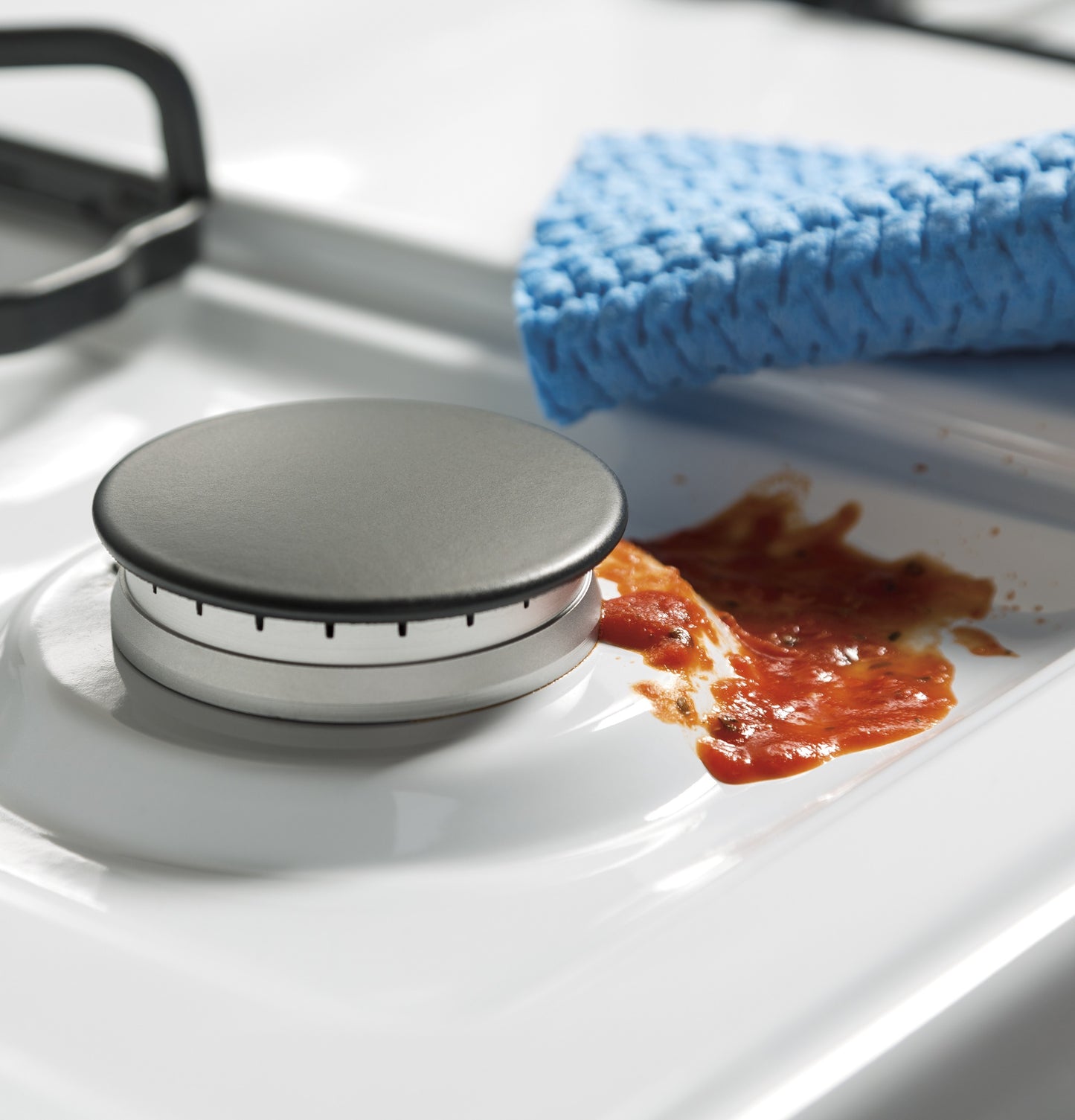Close-up of gas burner cap on white surface with spilled red sauce and blue cleaning cloth in kitchen range setting