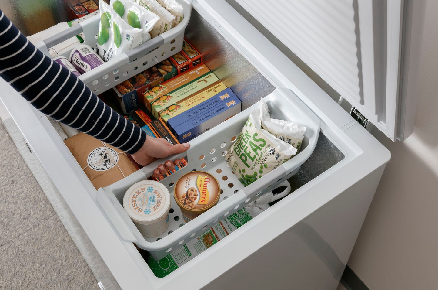 Open white chest freezer filled with frozen groceries and person removing plastic storage basket