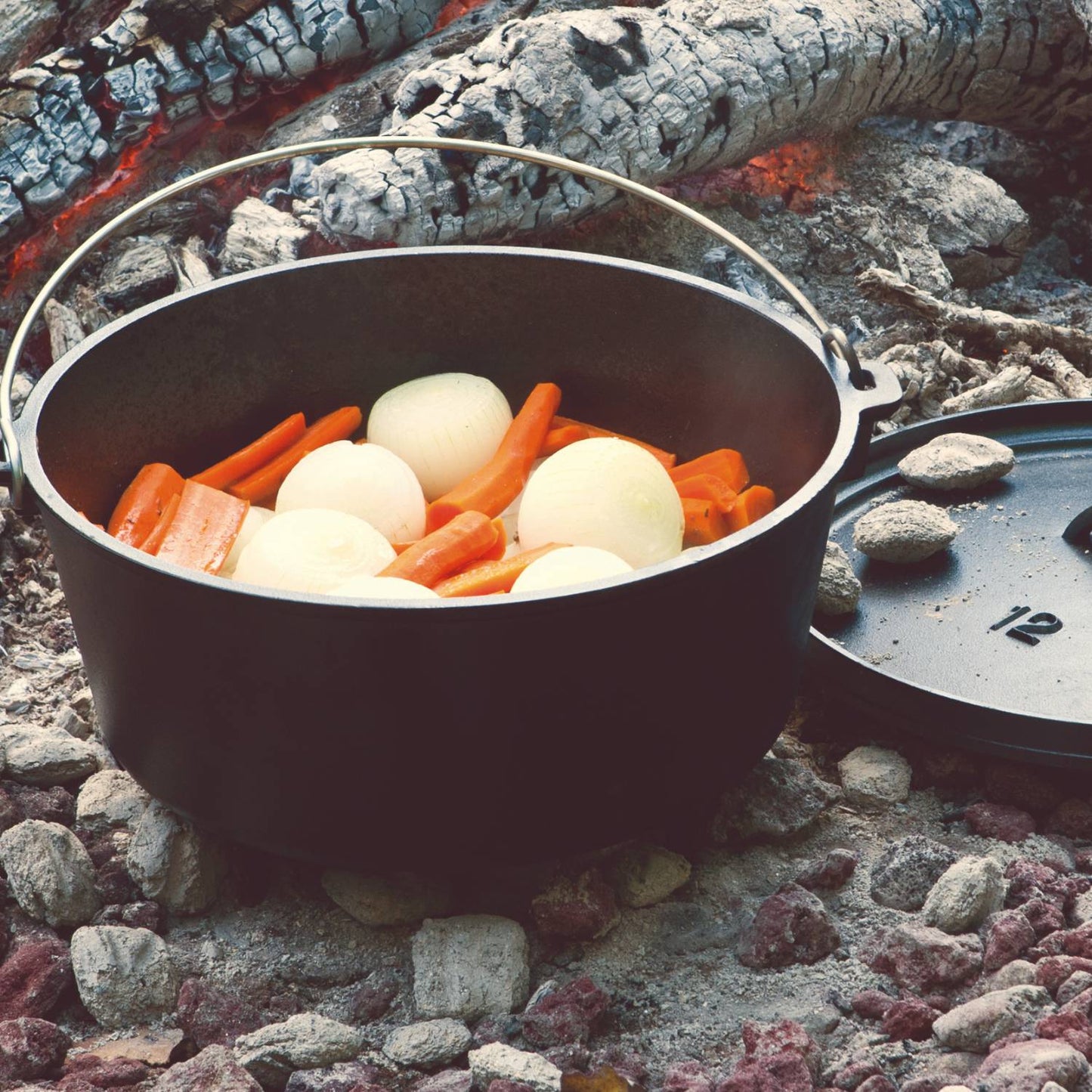 Camp Dutch oven with whole onions and carrots cooking over hot embers in kitchenware collection setting