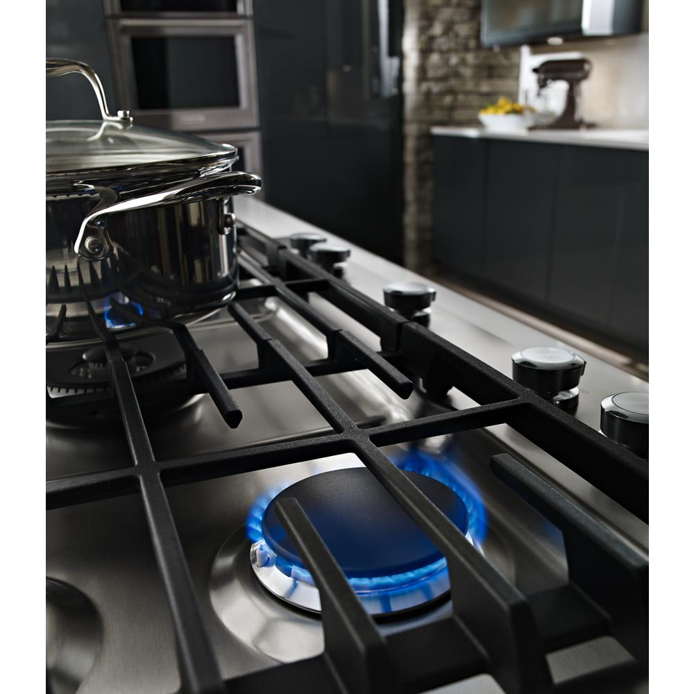 Blue flame burning on front burner of stainless steel 36-inch gas cooktop with black cast iron grates in kitchen setting