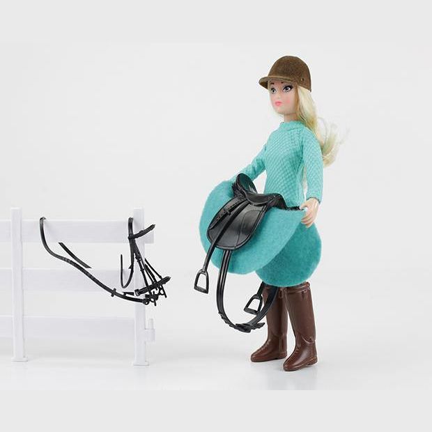 Doll dressed in teal riding outfit holding black saddle next to white fence with bridle accessory