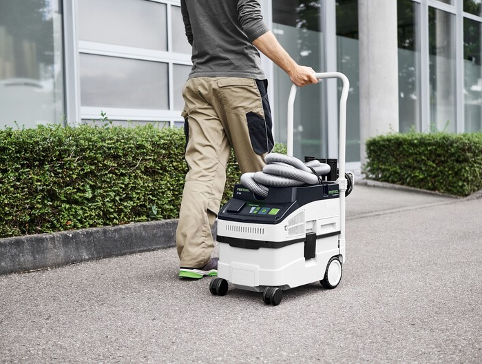 Person transporting a compact Festool dust extractor with hose and wheeled handle on outdoor pavement surface