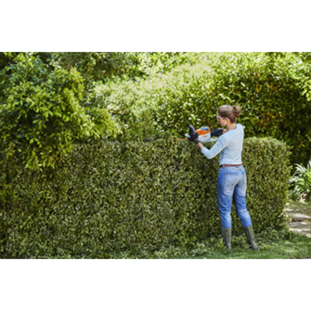 Woman trimming a thick green hedge outdoors using an orange and white cordless hedge trimmer with gloves and boots