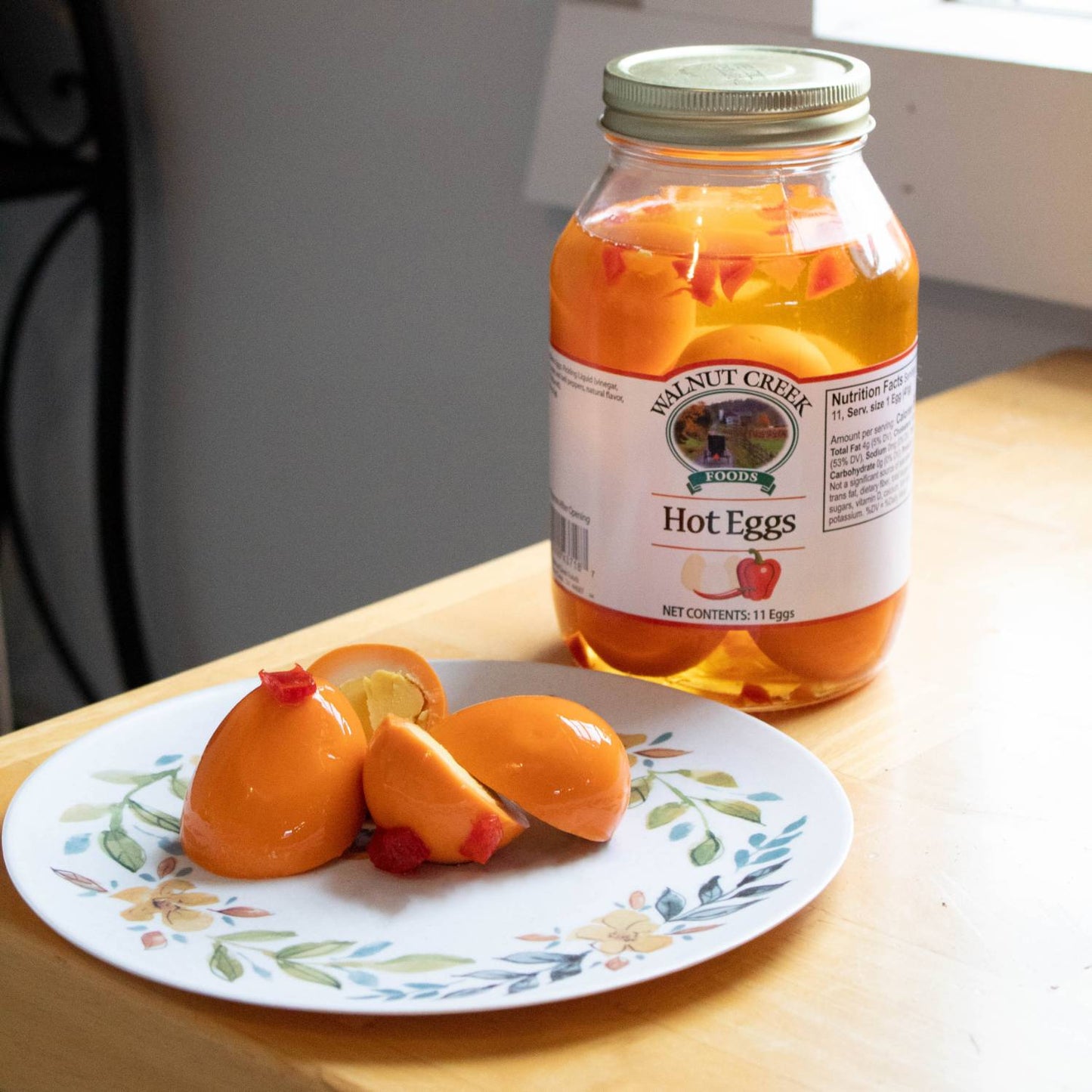 Glass jar of spicy pickled eggs with red chili pieces next to peeled eggs on floral plate for food and beverage.