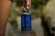 Person holding a blue insulated water bottle with a gray handle against a blurred natural outdoor background.