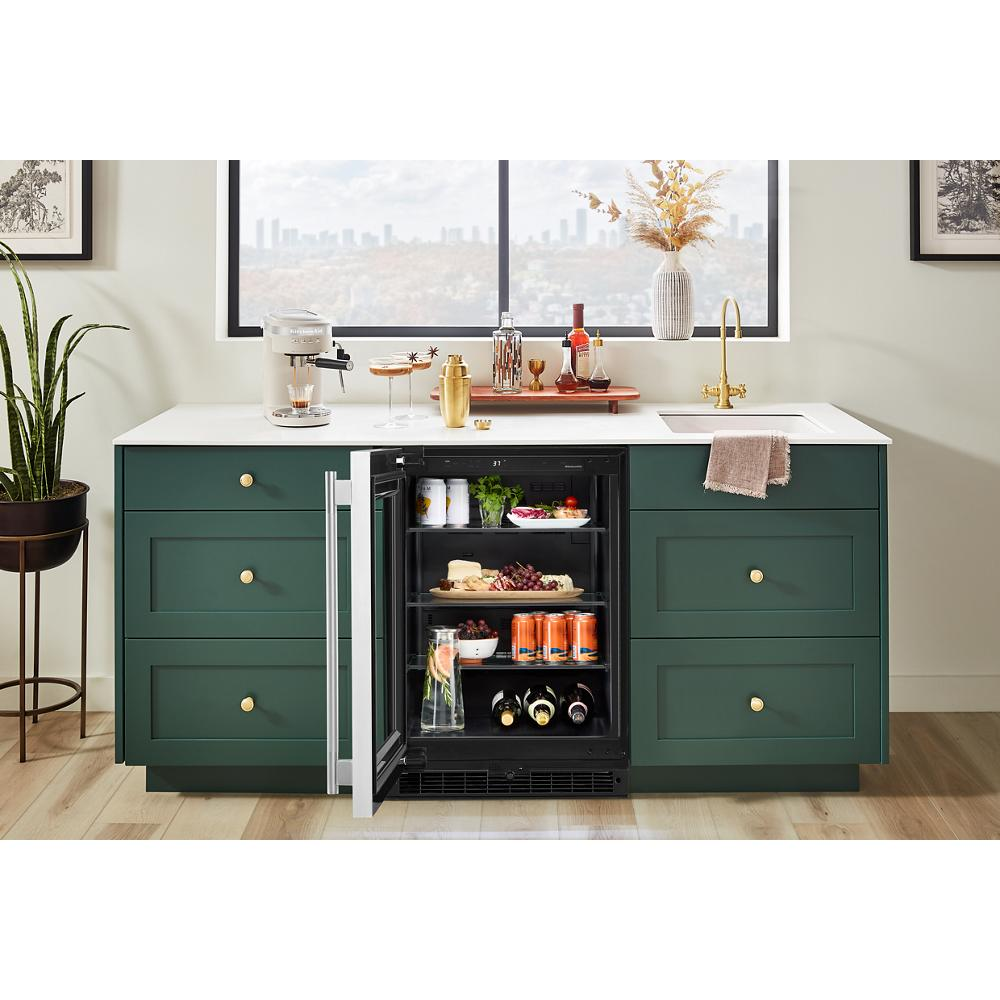 Undercounter refrigerator with glass door open, showing stocked shelves in green kitchen cabinetry setting with white countertop
