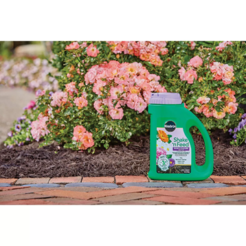 Green Miracle-Gro Shake 'n Feed container with purple lid placed by blooming rose bushes in outdoor garden setting