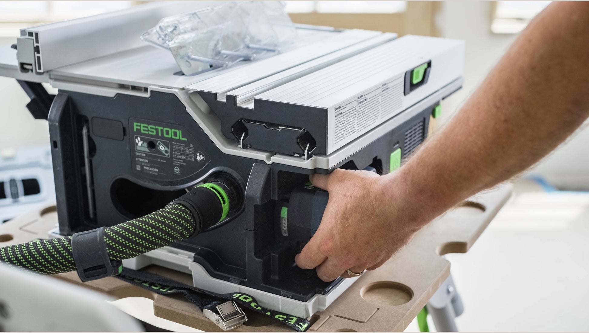 Hand operating the dust extraction port on a Festool cordless table saw with green and black hose attached, featured in Power Tools and Festool Cordless Saws collections.