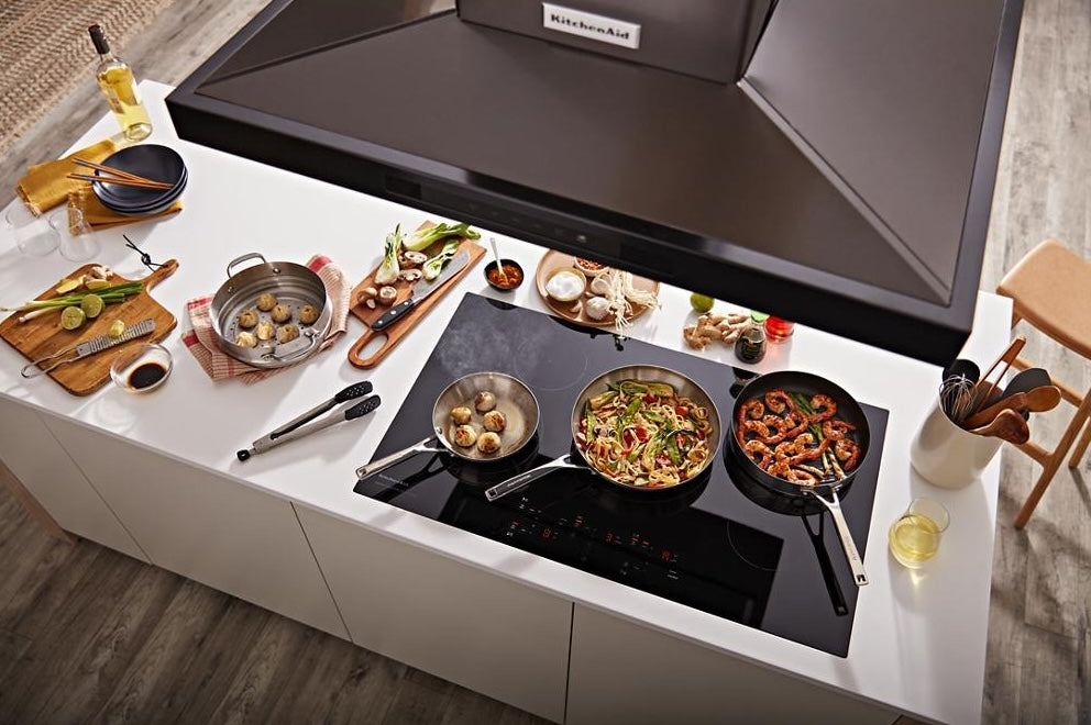 Black canopy hood mounted above a modern cooktop with three pans cooking various dishes on a bright kitchen island, part of Island Range Hoods and Kitchen & Kitchenware collections.