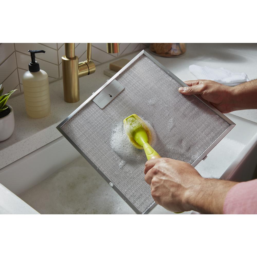 Hands scrubbing metal mesh filter with brush in kitchen sink, part of Island Range Hoods maintenance routine
