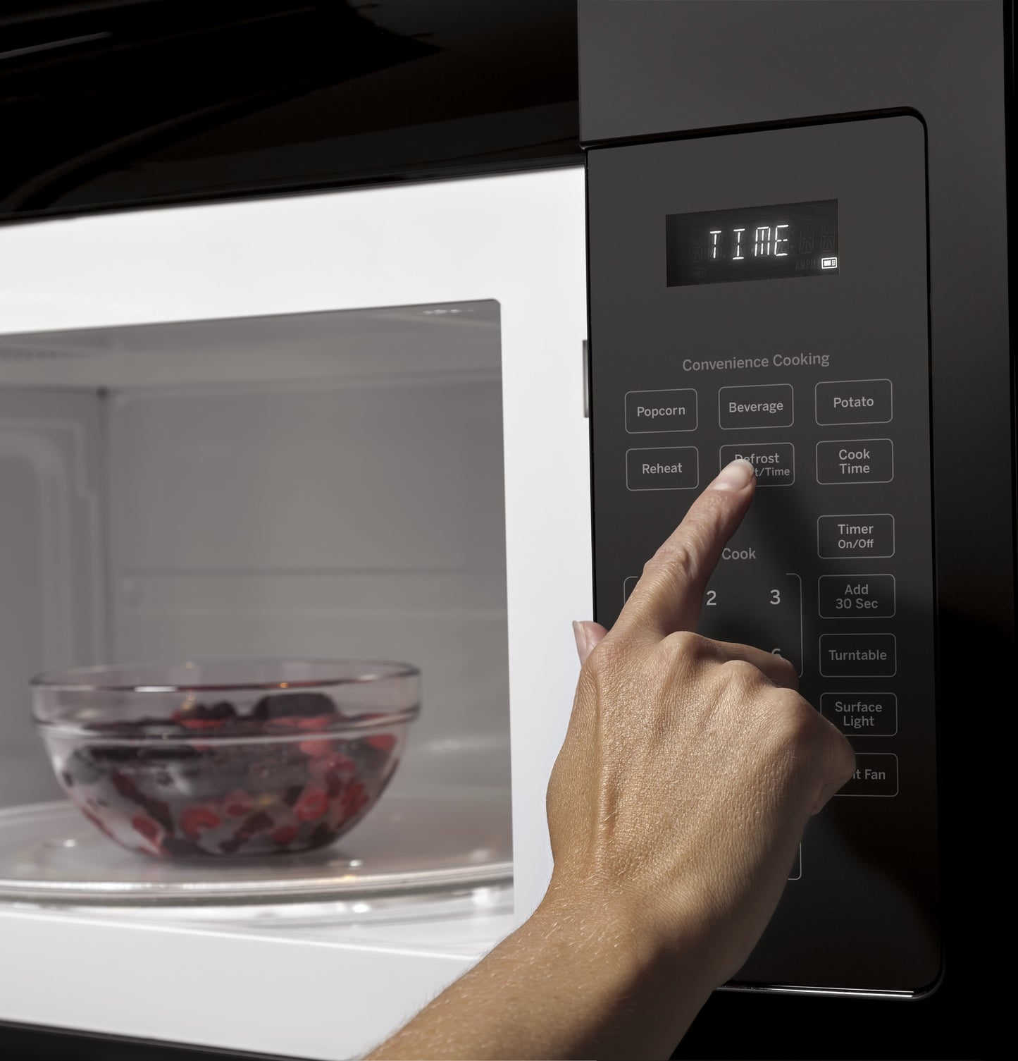 Finger pressing defrost option on black control panel of over-the-range microwave with glass bowl inside from kitchen appliances collection