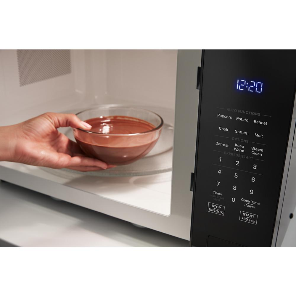 Hand placing glass bowl of food into sensor cooking microwave with digital control panel lit and active.
