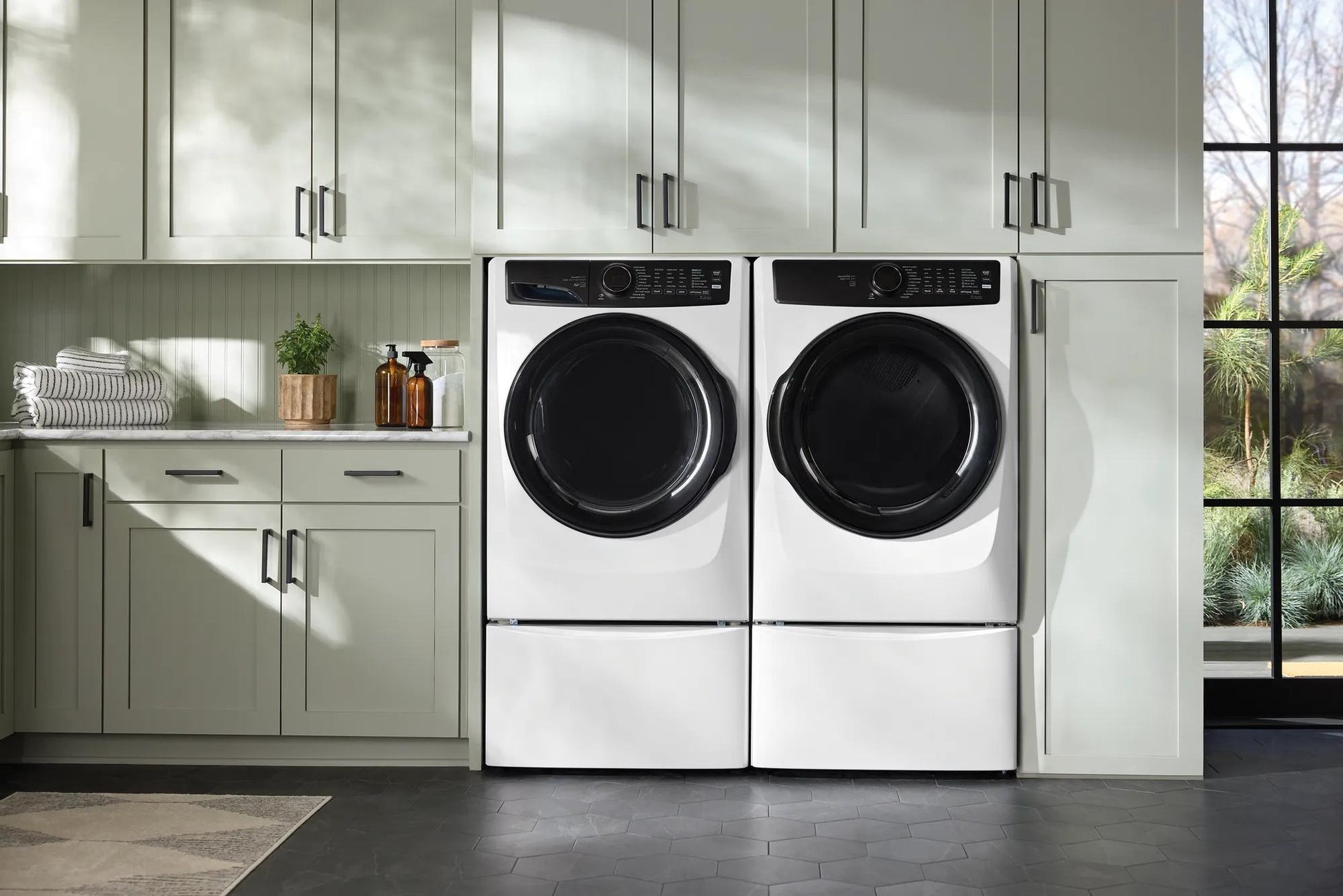 White front load washer and dryer with black doors in bright laundry room featuring gray cabinetry and natural light in appliances collection