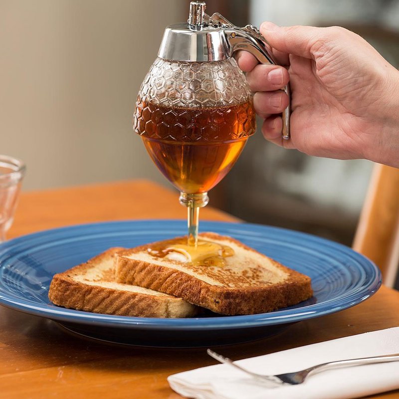Glass honeycomb-patterned syrup dispenser with metal lid pouring syrup over toasted bread on blue plate in kitchenware setting