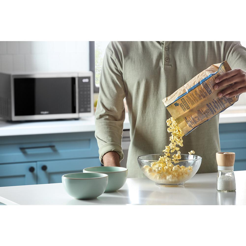 Man pouring popped popcorn into glass bowl on white countertop with stainless steel microwave in blue kitchen