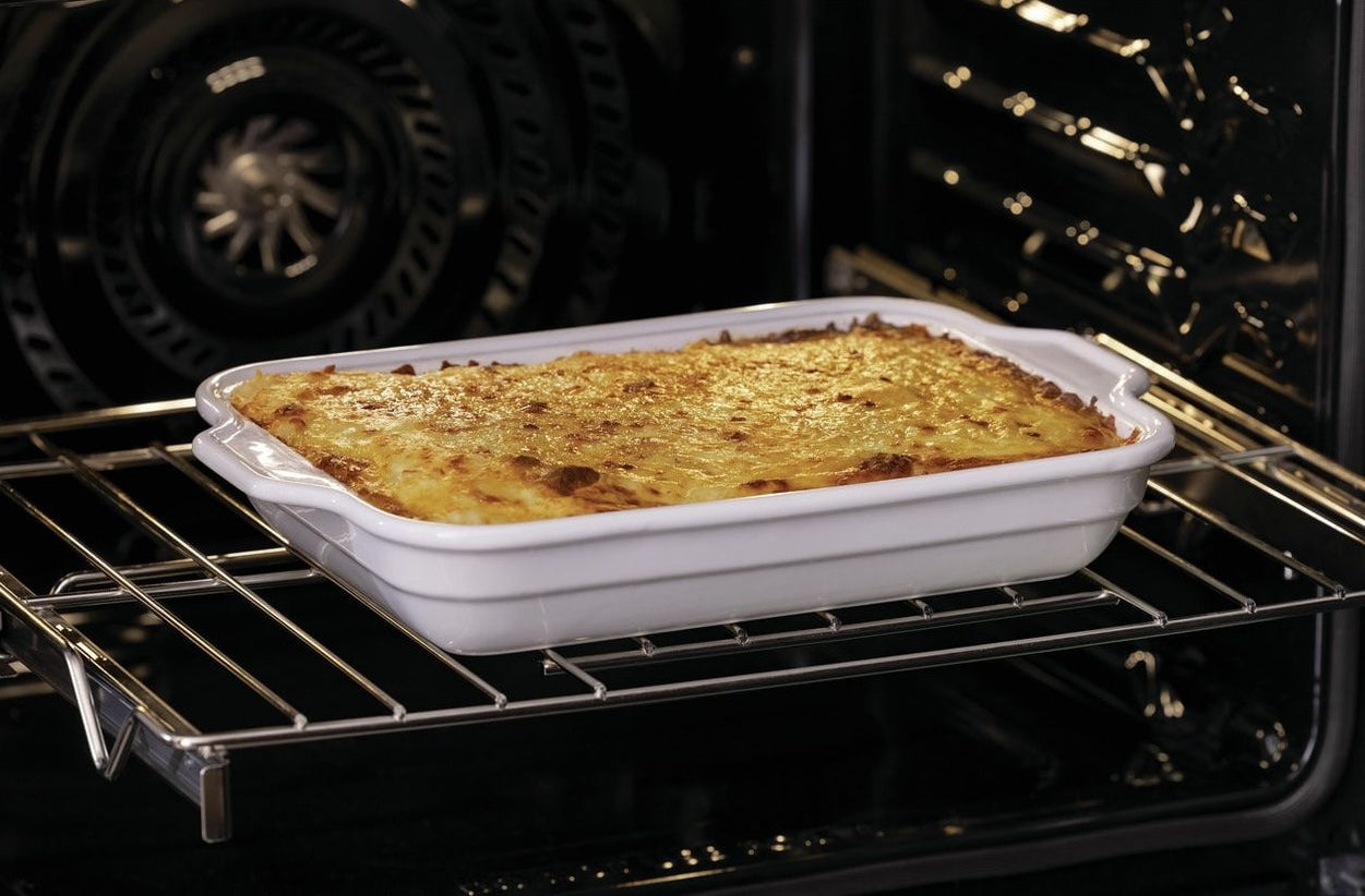 White ceramic dish with casserole baking on middle rack inside electric wall oven in kitchen appliances collection