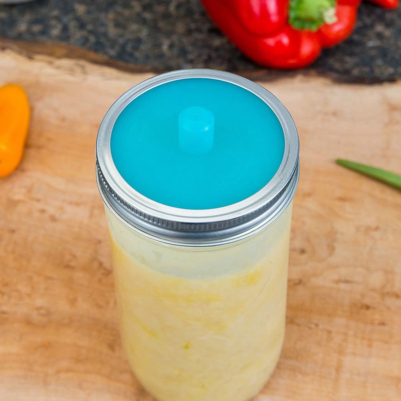Clear glass jar with blue fermentation lid filled with yellow fermenting vegetables on wooden board in kitchenware setting