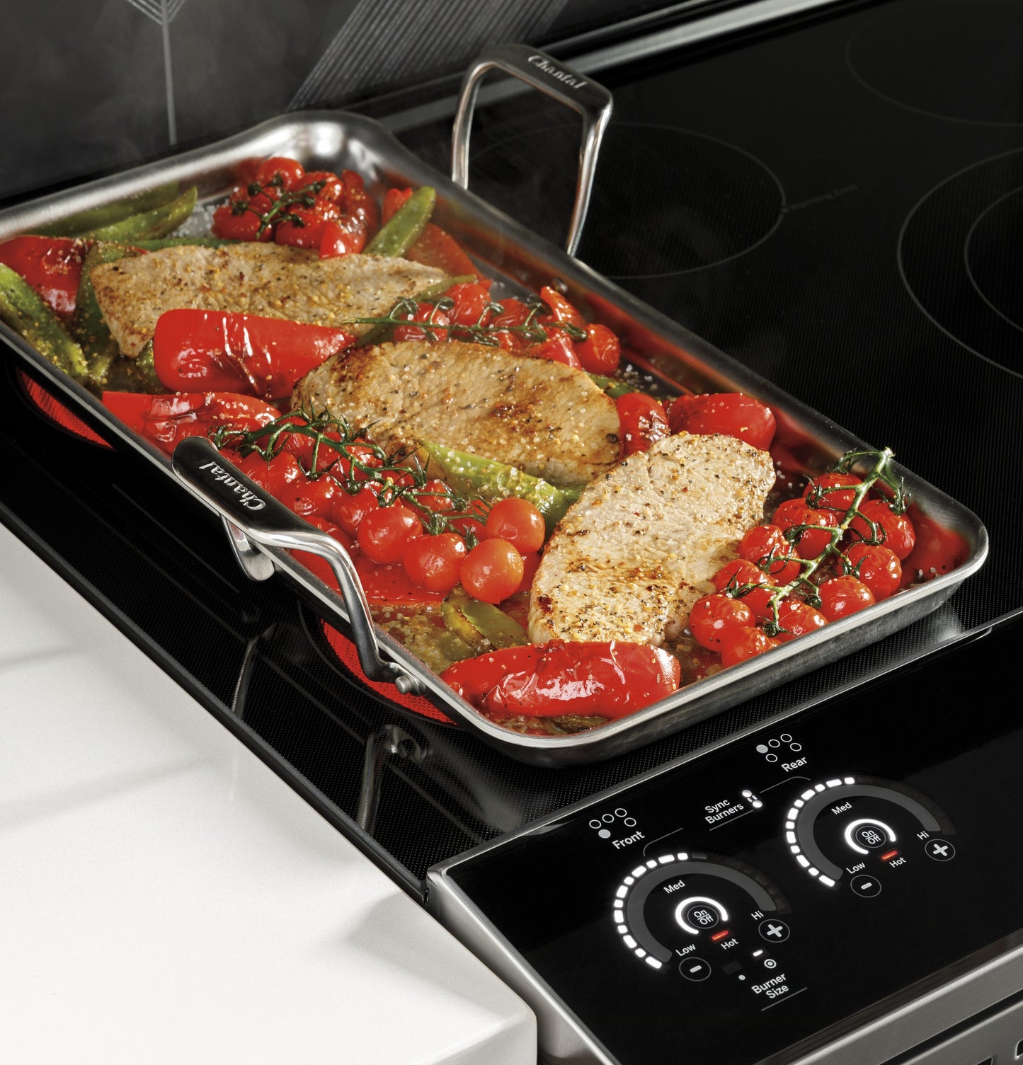 Stainless steel roasting pan with pork chops, cherry tomatoes, and vegetables on black induction cooktop controls