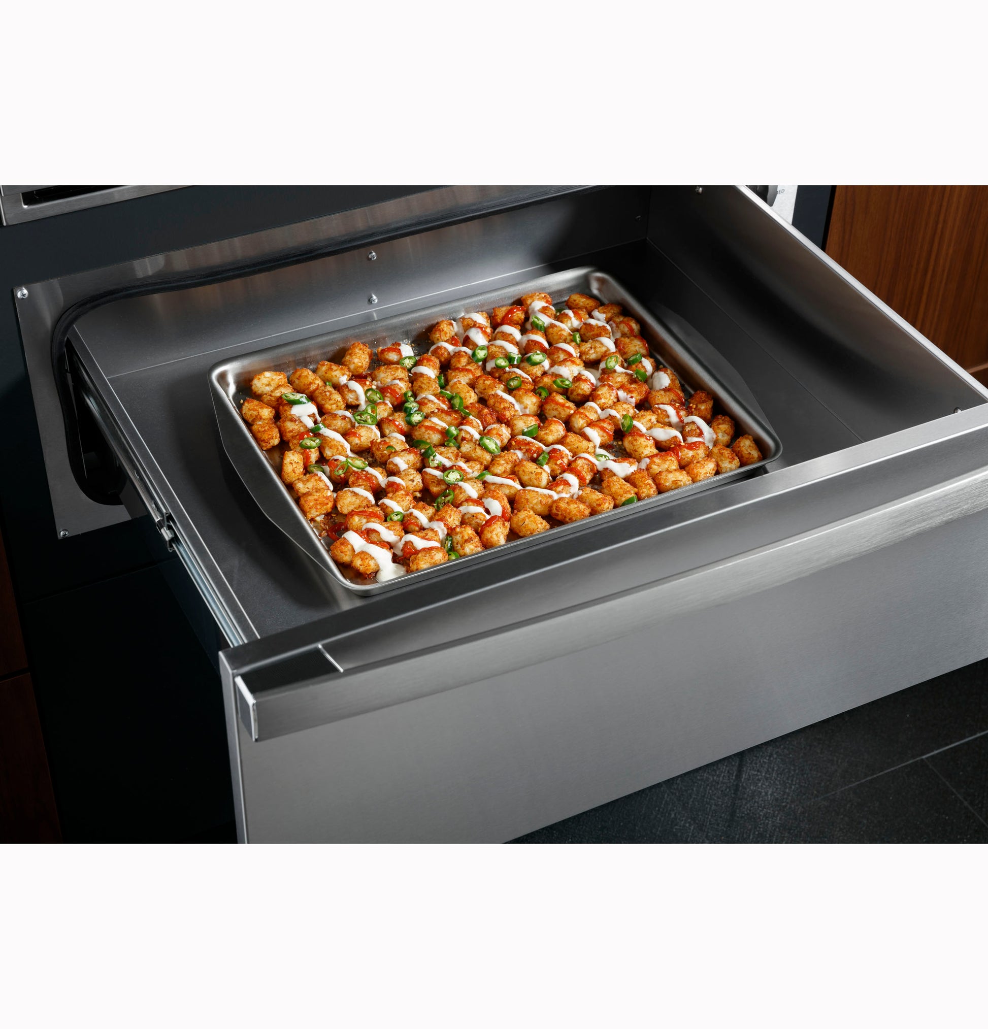 Stainless steel warming drawer open with a tray of seasoned tater tots, part of modern kitchen appliances collection.
