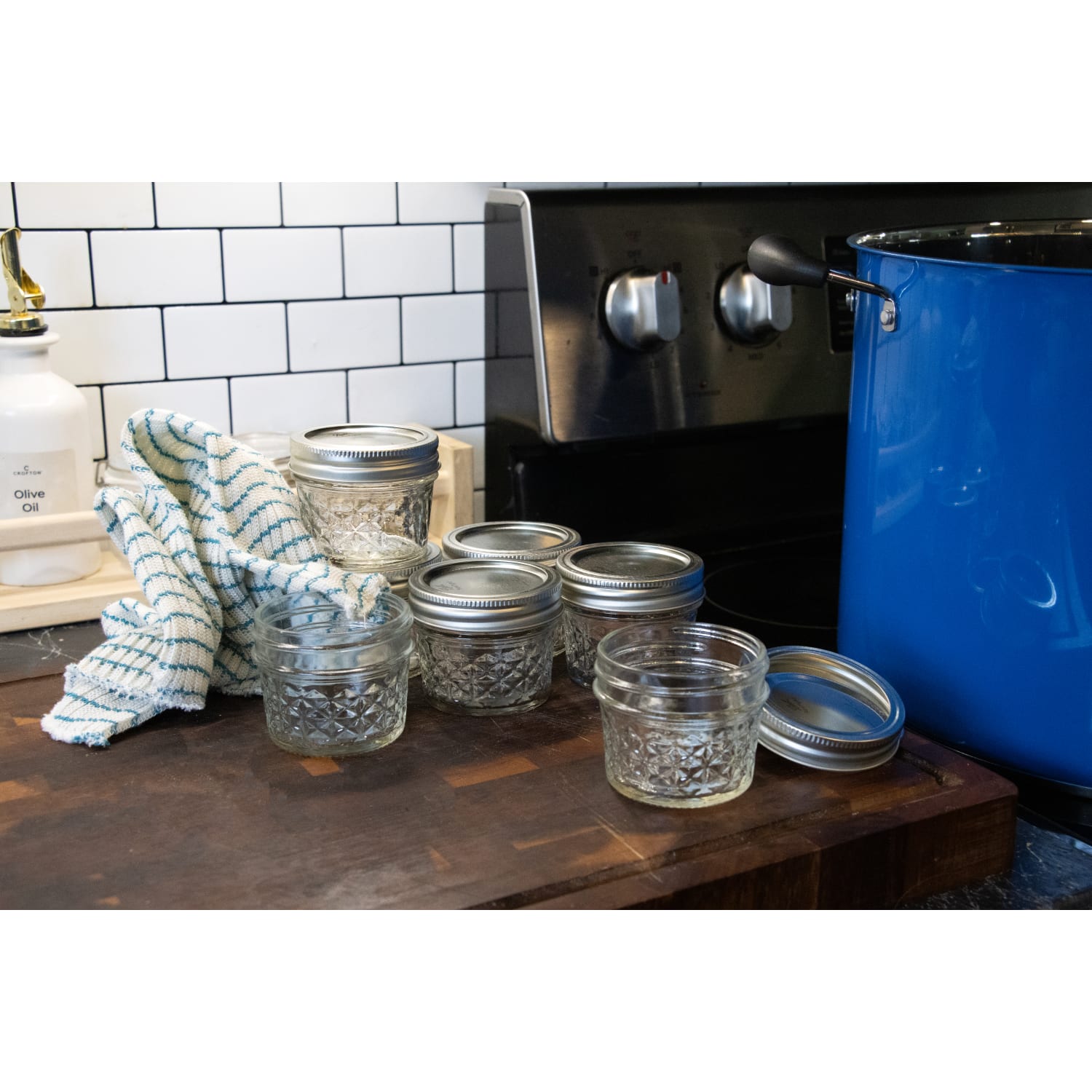 Clear small glass jars with metal lids arranged on wooden counter near blue pot and kitchen stove