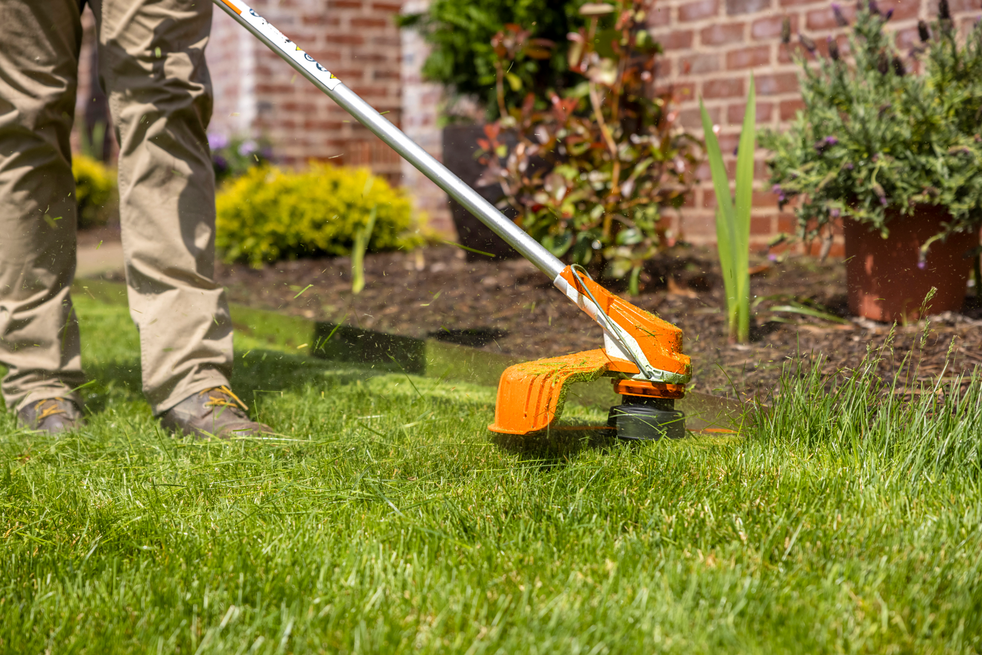 Person trimming lawn with orange and white battery-powered garden tool near flowerbed for home improvement tasks