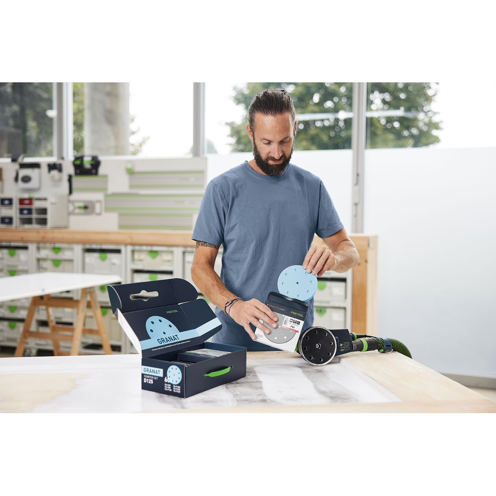 Man holding blue sanding discs near open box and green power sander in workshop with tool accessories and abrasives