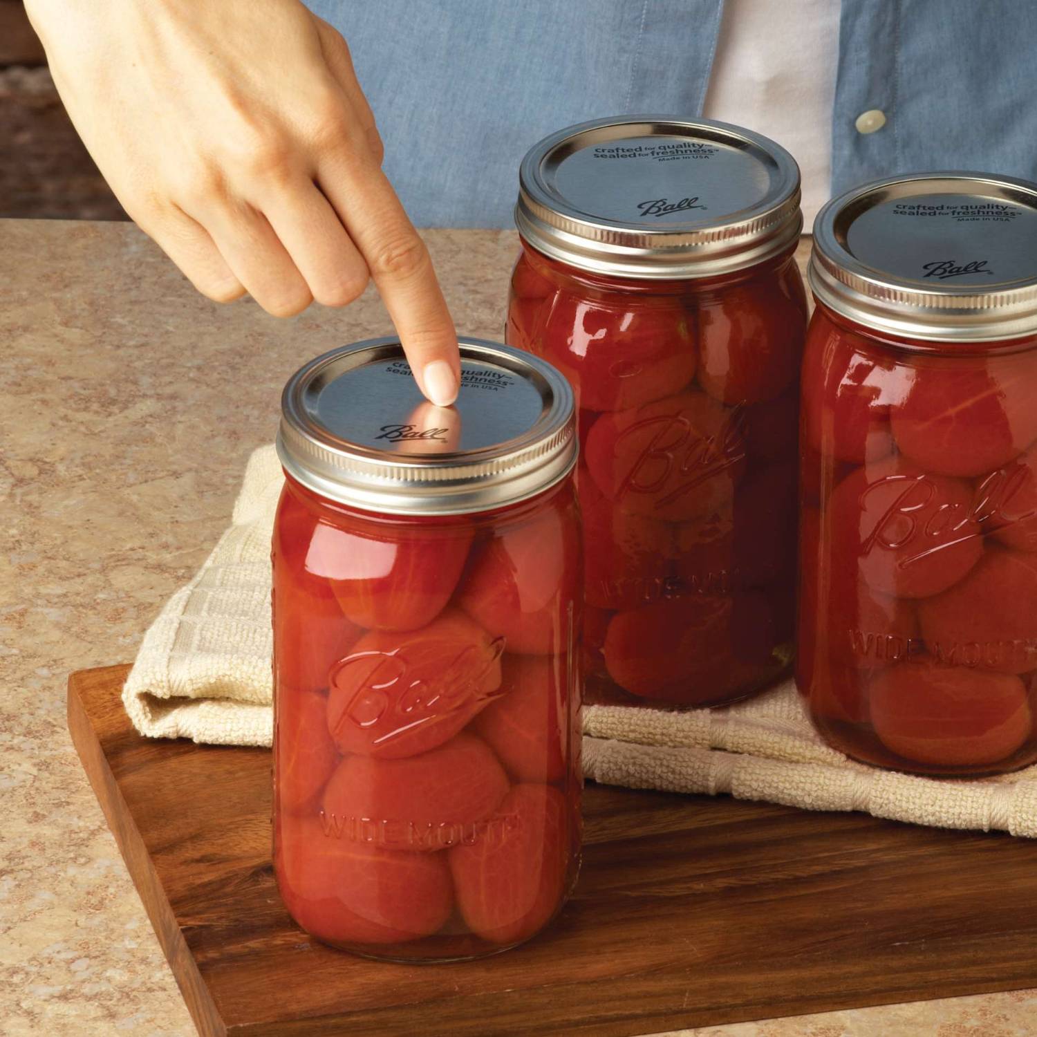 Hand pressing silver-toned metal dome canning lid on glass jar filled with preserved tomatoes on wooden board