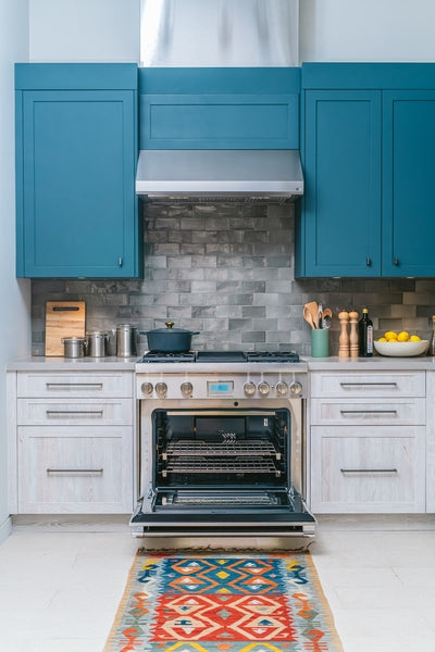 Open stainless steel dual fuel professional range oven surrounded by blue cabinetry and gray tiled backsplash in modern kitchen