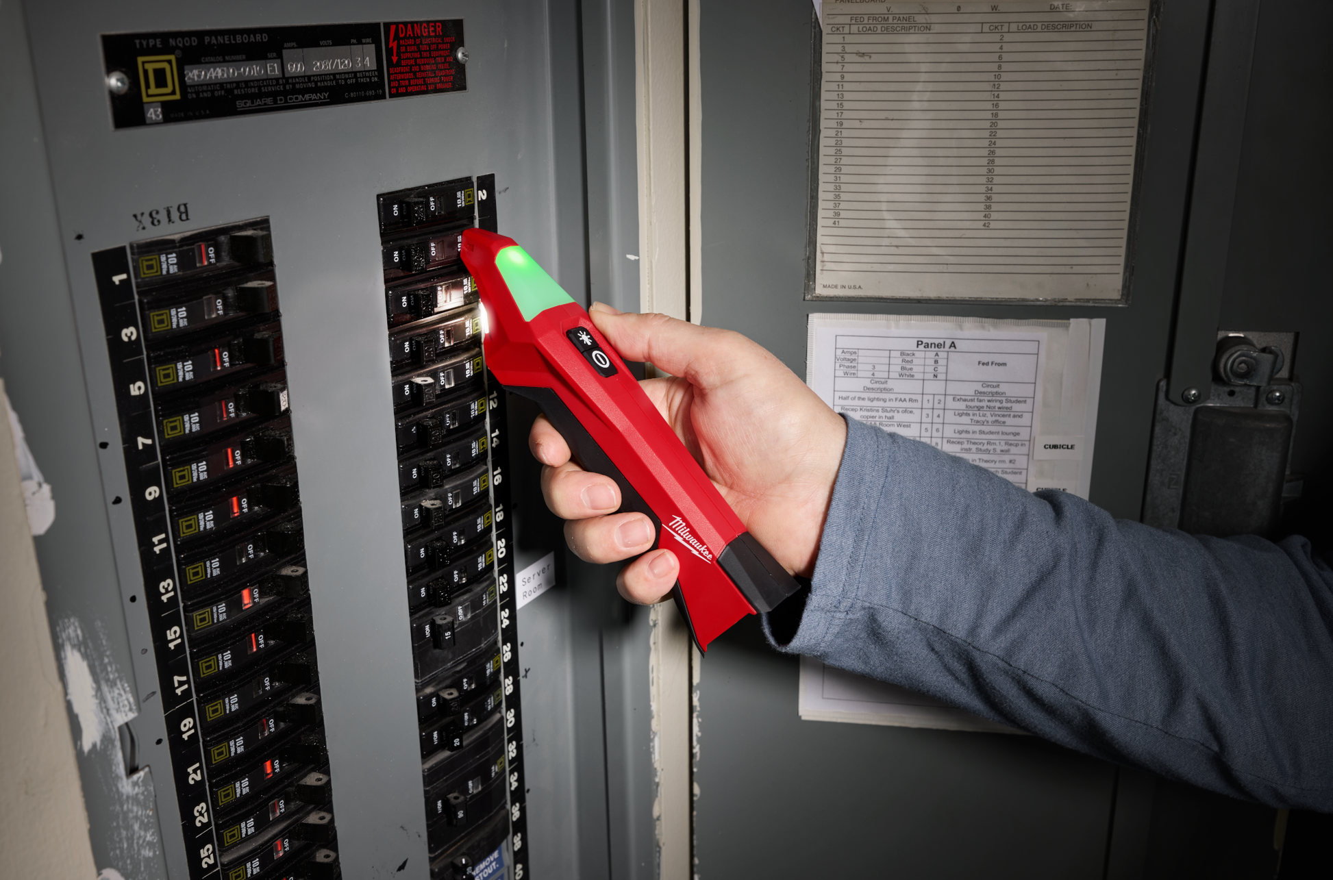 Hand holding red and black Milwaukee circuit breaker finder with LED light near electrical panel for home improvement